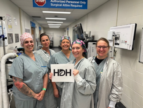 Five healthcare workers in surgical scrubs and caps posing together in hospital corridor, one holding HDH sign